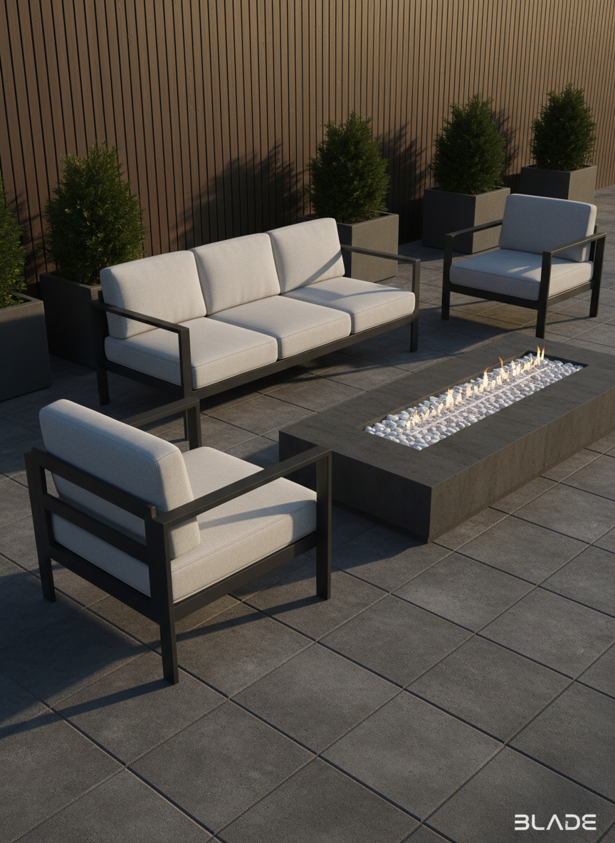 A refined outdoor seating area featuring low-profile, matte black metal lounge furniture with pale stone-colored cushions, set atop large-format charcoal pavers. The arrangement is precisely positioned beside an angular, linear gas fire pit with a trough of smooth white river stones. Minimalist evergreen shrubs in geometric planters line the rear, extending along a privacy wall clad in vertical dark wood slats. Late afternoon sunlight casts long, soft shadows and gentle highlights across the textured pavers and cushions, enhancing tactile details. The scene is shot from a slightly elevated, three-quarters angle, balancing warmth and formality with cinematic clarity and a subdued, harmonious palette. The composition and mood communicate understated sophistication, ideal for showcasing BLADE’s commitment to elevated outdoor living spaces.